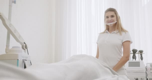 Ung flicka med blont hår läkare kosmetolog tar bort mask leende och sätter händerna i en kosmetisk ljusa kontor — Stockvideo