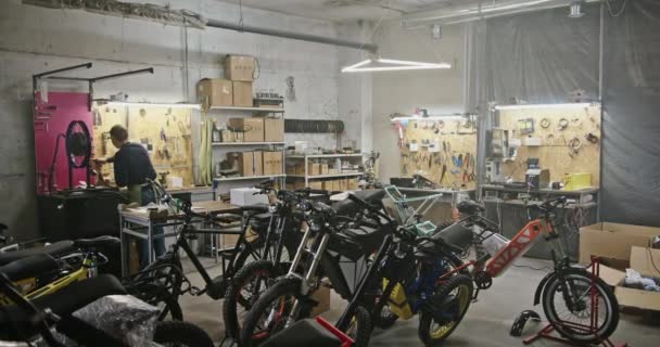 Proyek eco bisnis lokakarya besar untuk produksi sepeda listrik. Seorang pria bekerja pada latar belakang berbagai macam sepeda listrik sepeda eco sepeda di sebuah ruangan besar dengan pencahayaan buatan — Stok Video