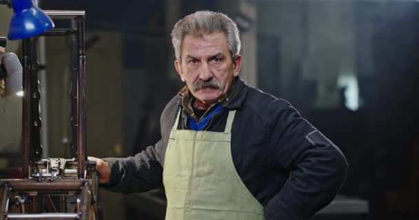 Mecánico viejo cansado limpiando el sudor. Hombre mayor agotado con bigote limpiando el sudor de la frente y sonriendo para la cámara y luego empezando a reparar el mecanismo de metal en el taller — Vídeos de Stock