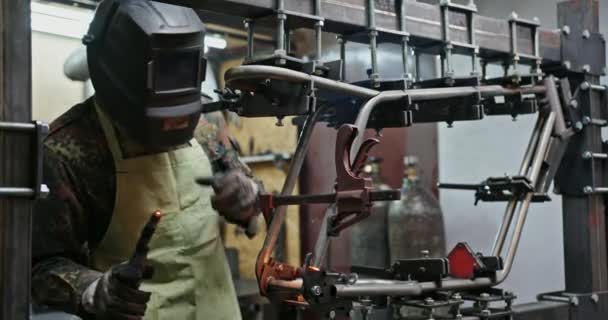 Een jongeman lassen ijzeren onderdelen stopt en kijkt naar het lassen blijft zijn werk in een beschermend pak met handschoenen en een lasmasker in zijn werkplaats — Stockvideo