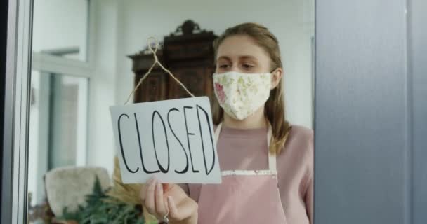 Una joven que es dueña de un pequeño negocio vestido con una máscara entrega un letrero abiertamente y abre su floristería durante la cuarentena y el virus — Vídeo de stock