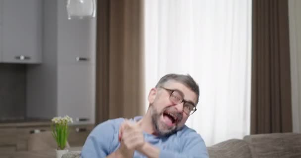 Middle aged fan celebrating victory on sofa. Mature man gesticulating and screaming while sitting on couch and celebrating victory in match — Stock Video