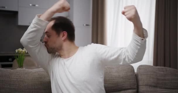 Stærk europæisk mand viser biceps kysser dem, der viser sin styrke med hænderne sidder på sofaen derhjemme på baggrund af et lyst køkken – Stock-video