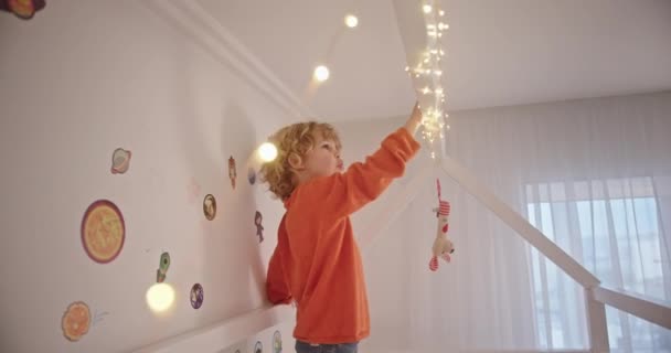 Ternopil, Ucrânia, 6 de outubro de 2020: Little curly boy standing on the bed in a cozy bright childrens room playing and hanging a garland dreaming laughs happy child — Vídeo de Stock