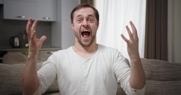 Man rejoices shouting with happiness victory raises hands looks at camera sitting on sofa at home in modern apartment — ストック動画