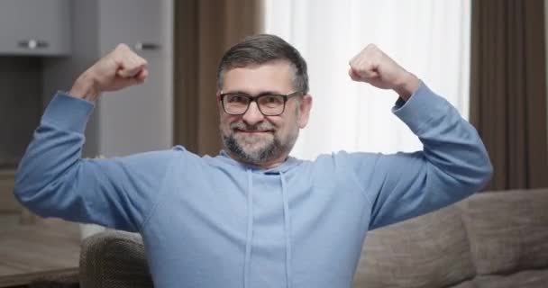 L'uomo sorridente mostra la sua forza con le mani. Forte vecchio con la barba mostra le mani bicipiti mentre si siede a casa sul divano — Video Stock