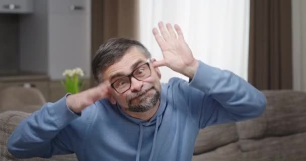 Hombre maduro tonto haciendo caras. Divertido hombre de mediana edad en gafas mirando a la cámara y haciendo muecas mientras está sentado en el sofá en casa — Vídeo de stock