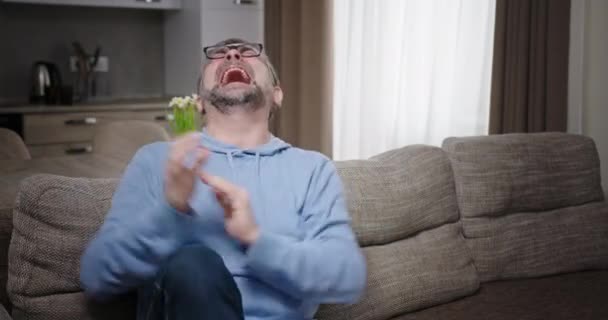 Mature man celebrating success on sofa. Middle aged male in glasses clenching fists and laughing loudly while sitting on couch and celebrating success at home — Stock Video