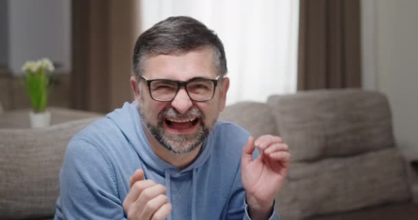 Старший мужчина в очках эмоционально смеется хлопает в ладоши показывает, как слушает шутку, сидя дома на диване в яркой квартире — стоковое видео