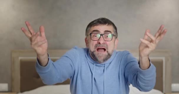 Angry man screaming at camera. Enraged mature male in glasses gesticulating and yelling at camera in anger while sitting on bed at home — Stock Video