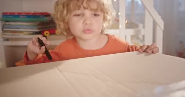 Lindo dibujo de niño en caja de cartón. Tracking shot de adorable chico con pelo rizado hablando y dibujando en caja de cartón en casa — Vídeo de stock