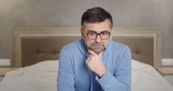 Серьезный взгляд на камеру старший мужчина расстроен из-за плохих новостей безэмоциональная депрессия сидит дома на кровати в спальне — стоковое видео