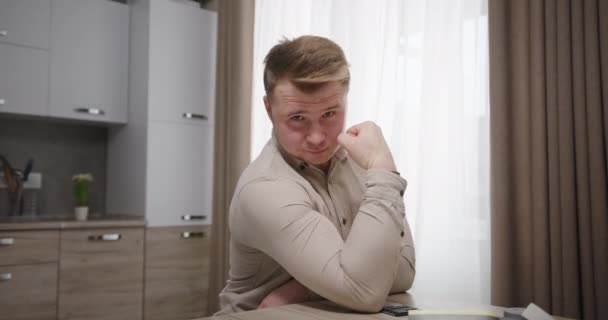 Confident man showing biceps in kitchen. Self assured male demonstrating muscles then crossing arms and smirking while sitting at table in kitchen at home — Stock Video