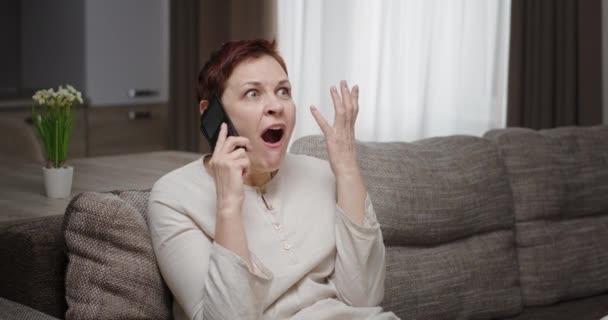 Conversa positiva chocante alegre no telefone. Mulher falando ao telefone em casa sentado no sofá rindo surpreso — Vídeo de Stock