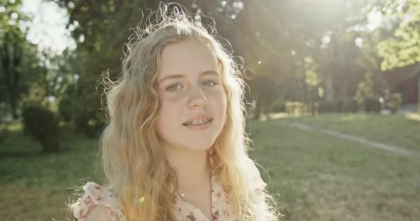 Porträt der schönen Lockenkopf mit Zahnspange in der strahlenden Sonne im Sommerpark vor dem Hintergrund der Bäume. — Stockvideo