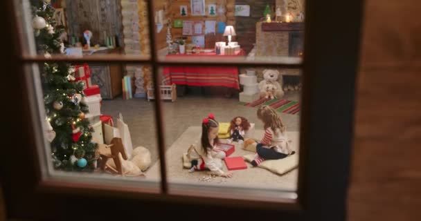 Dvě holčičky si večer před Vánocemi hrají na koberečku s panenkami v pohádkovém domku u krbu a čekají na vánoční svátky. Pohled oknem z okna — Stock video
