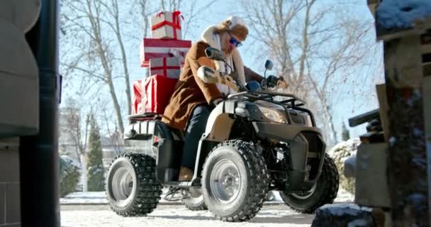 Papá Noel moderno. Alegre Santa Claus entrega regalos en un ATV en el día de Navidad en el fondo de una casa de nieve el fondo. — Vídeos de Stock