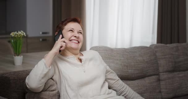 Αστεία ώριμη γυναίκα κουτσομπολεύει με φίλο στο smartphone. Ευτυχισμένη μεσήλικη γυναίκα με κοντά κόκκινα μαλλιά κάνει αστεία πρόσωπα και να μοιράζονται τα νέα με το φίλο στο κινητό, ενώ κάθεται στον καναπέ στο σπίτι — Αρχείο Βίντεο