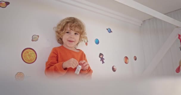 Маленький кудрявый блондин улыбается, смеясь, пытаясь открыть жевательную резинку конфеты, стоящие в детской на кровати на фоне рисунков — стоковое видео