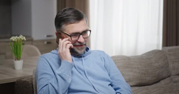 Człowiek rozmawiający zabawnie przez telefon uśmiechnięty zaskoczony siedzeniem w domu na sofie nowoczesne wnętrze. Rozmowa telefoniczna jest szokująco pozytywna. — Wideo stockowe