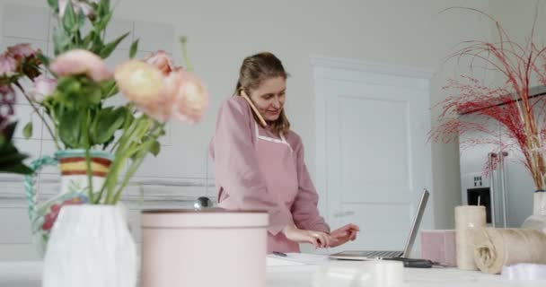 Młoda sprzedawczyni dziewczyna w kwiaciarni kwiaciarnia biznes pani mówi przez telefon uśmiechając się i pisząc zamówienie w laptopie stoi za ladą — Wideo stockowe