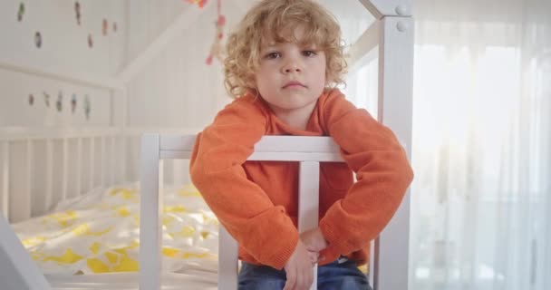 Pequeño niño feliz y emotivo pensativo con el pelo rubio rizado sentado en la cama colgando los brazos hacia abajo pensando en algo — Vídeos de Stock