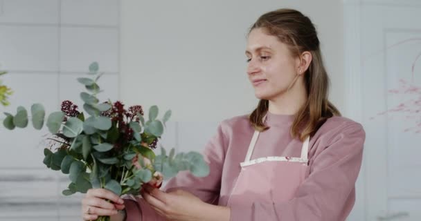 Junges Blumenmädchen kreiert einen Strauß frischer Blumen als Geschenk für den Käufer lächelt liebt seinen Job glücklich blickt in die Kamera in einer Schürze — Stockvideo