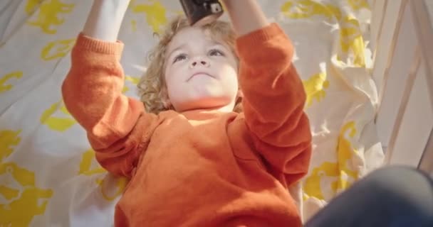 Pojken leker med ficklampan på sängen. Från ovan söt pojke i casual kläder leka med glödande ficklampa när du ligger på sängen hemma — Stockvideo