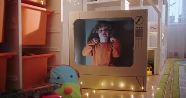 Kis szőke göndör fiú ül egy kartondobozban karton TV zseblámpák álmok és feltalál egy fantasy játék. Gyermekbarát babaszoba. — Stock videók