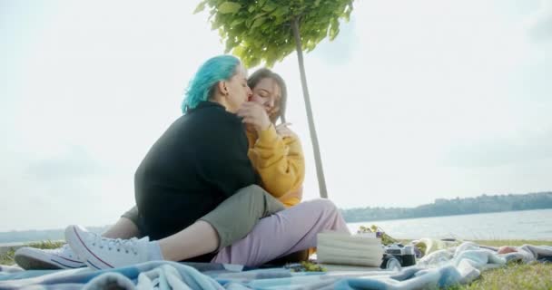 Leszbikus pár ölelkezik piknik közben a folyó közelében. Földszint fiatal barátnők ölelik egymást, miközben pihennek a takarón piknik közben a folyó partján hétvégén — Stock videók