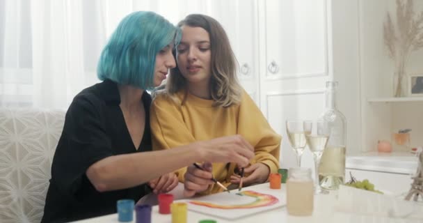 Two young lesbian women painting the Gay Pride rainbow symbol together in their apartment while celebrating with a bottle of wine — Stock Video