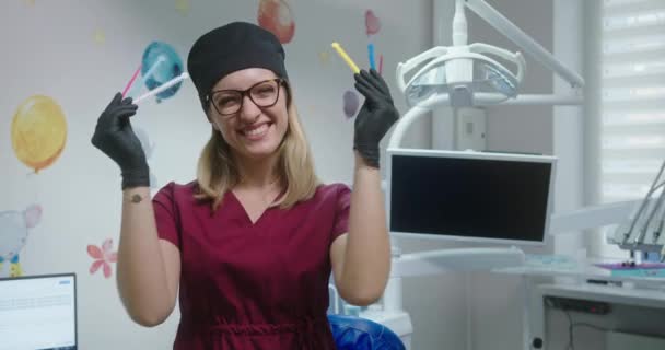 Jonge vrouwelijke arts tandarts orthodontist met gekleurde ligatuur rubberen band voor beugels lachen stijgers in de tandartspraktijk in een dokters jurk en hoed — Stockvideo