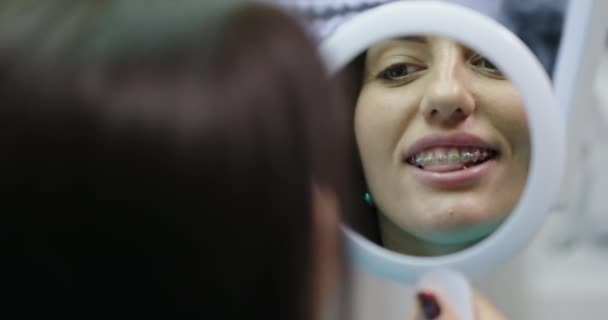 Paciente jovem na recepção do dentista ortodontista olha no espelho para os dentes com aparelho sorri feliz com o resultado feliz — Vídeo de Stock