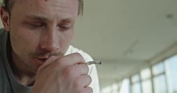 Mladý muž je sklíčený apatií poté, co závislost na alkoholu sedí v jasně opuštěném pokoji, kouří cigaretu a přemýšlí — Stock video