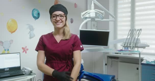 Jonge vrouwelijke tandarts orthodontist met bril glimlacht en zet zijn handen staande in de kinderen tandheelkundige kantoor in een artsen badjas hoed en handschoenen — Stockvideo