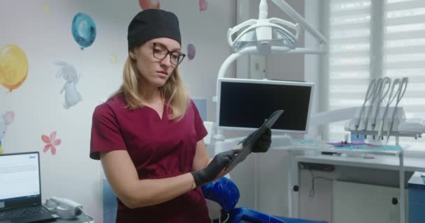 Молодая женщина-стоматолог Профессионал, стоящая в своей клинике с фотографией зубов пациента, смотрящего на фотографию, решает процесс лечения, глядя в камеру и улыбаясь в медицинской одежде — стоковое видео