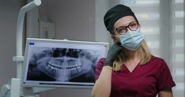 Młoda dentystka dentystka patrzy w kamerę poważnie zdejmuje maskę i wkłada ręce do okularów szlafrok i kapelusz — Wideo stockowe