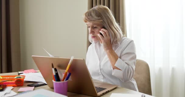 Covid-19 salgını sırasında evde çalışan yaşlı bir kadın dizüstü bilgisayarının başında oturan bir müşteri ya da meslektaşıyla evrak işlerini tartışıyor. — Stok video