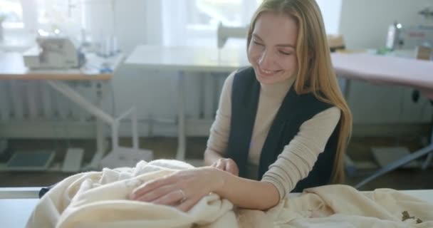 Gelukkige naaister die borduurwerk op stof onderzoekt. Hoge hoek van vrolijke vrouw glimlachend en bewonderende delicate plant ornament op jurk terwijl zitten aan tafel in licht atelier — Stockvideo