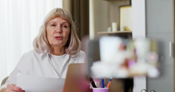 Senior teacher recording online lesson. Rack focus of elderly woman with paper speaking with online students while recording video for educational blog — Stock Video