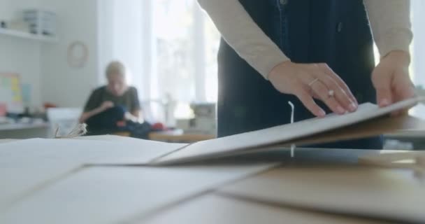 Αγνώριστη μοδίστρα συλλέγει σκίτσα στο τραπέζι. Επίκεντρο της ανωνυμίας γυρίζοντας σελίδες από μπλοκ ζωγραφικής μετά συλλέγοντας φύλλα χαρτιού με σχέδια από πάγκο εργασίας και φεύγοντας κατά τη διάρκεια της εργασίας — Αρχείο Βίντεο