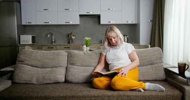 Mulher sênior com bebida quente e livro descansando no sofá — Vídeo de Stock