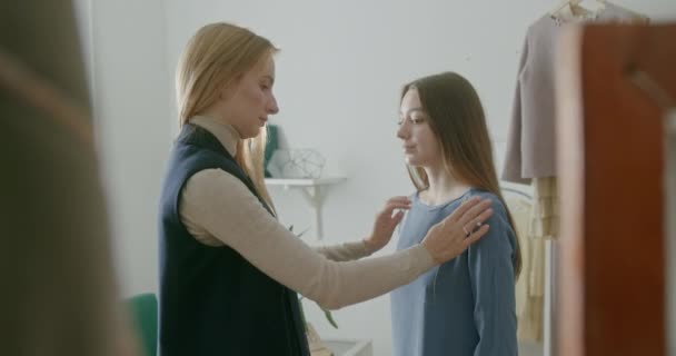 Female tailor adjusting dress with embroidery on client. Blond woman adjusting custom blue dress with floral embroidery on young female customer while working in studio — Stock Video