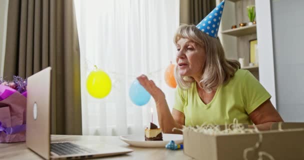 Elderly woman celebrating birthday online. Senior woman speaking with online friends and blowing candle on piece of cake during remote birthday celebration at home — Stock Video
