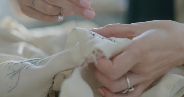 Oříznout švadleny šití na tkanině v dílně. Detailní anonymní žena s jehlou a nití šití květinové ornament na bavlněné šaty při práci v ateliéru — Stock video