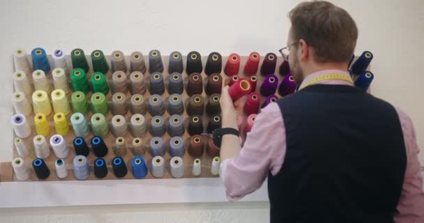 Портной выбирает правильную цветовую нить для шитья своей одежды из настенной стойки катушек или катушек в цветах спектра — стоковое видео