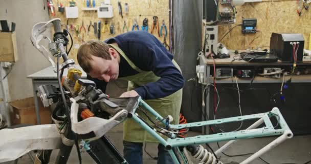 Człowiek pracuje nad stworzeniem metalowej ramy dla roweru elektrycznego, sprawdza te szczegóły, znużony wycieraniem czoła ręką w warsztacie i specjalnymi ubraniami — Wideo stockowe