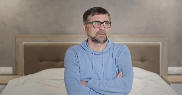Homem sênior com cabelos grisalhos em óculos ofendidos coloca suas mãos rola seus olhos não quer ouvir caretas sentadas na cama em casa — Vídeo de Stock