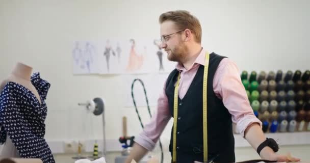 男性デザイナーはマネキンの近くの彼のスタジオに立って、彼が作成したドレスを調べ、カメラを見て、専門的に笑顔を見せ、手を置く — ストック動画