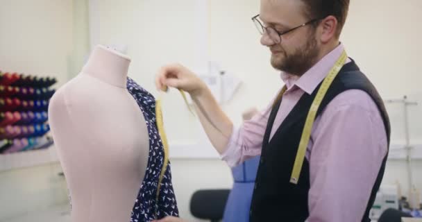 Hombre joven diseñador elegante crea un nuevo vestido mide los centímetros de cinta métrica de los parámetros de la tela en un maniquí de pie en su taller — Vídeos de Stock